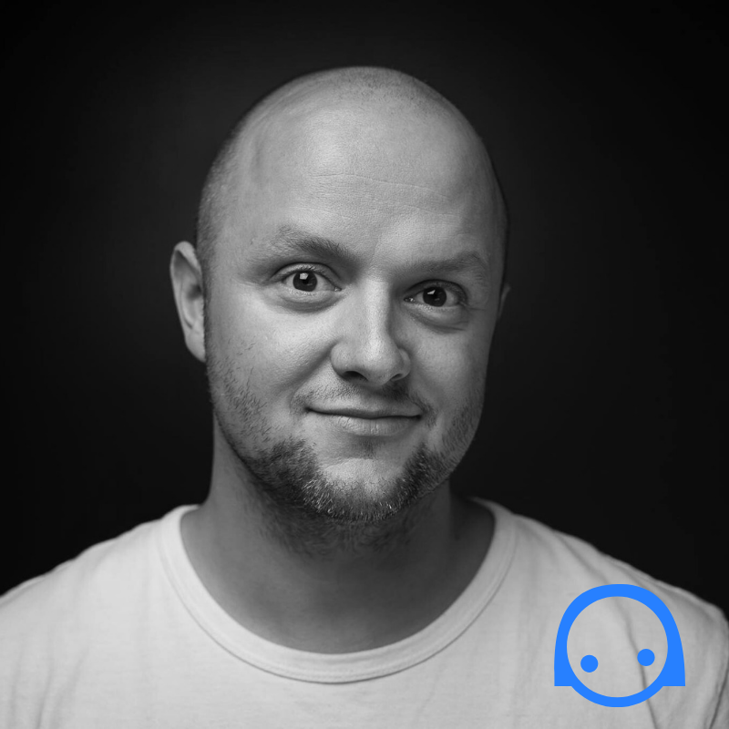 Hymyilevä kalju mies valkoisessa t-paidassa poseeraa tummaa taustaa vasten. Kuvan oikeassa alakulmassa näkyy yksinkertainen sininen logo, joka muistuttaa hymyilevää kasvoa.